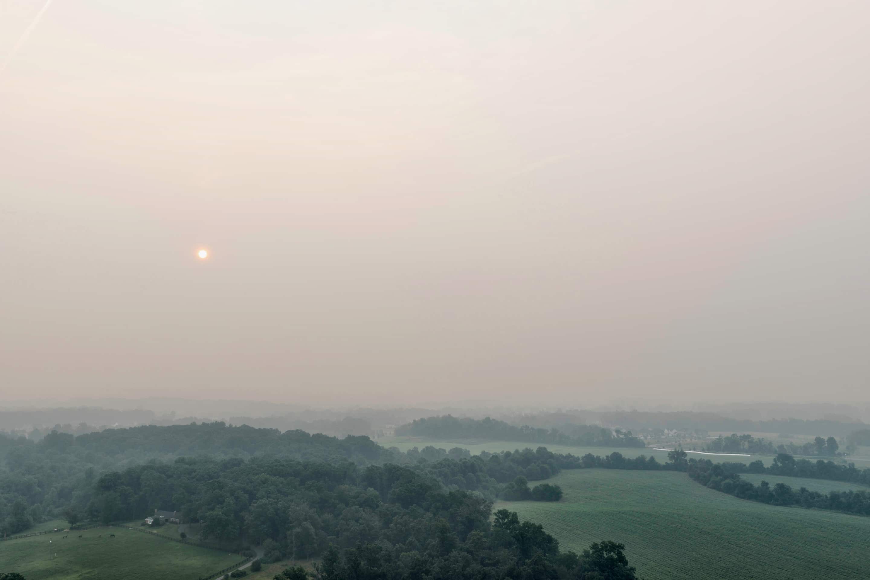 Wildfire smoke flattening a whole river valley into pale grey, with the sun reduced to a small orange disc above ordinary farmland and woodland.